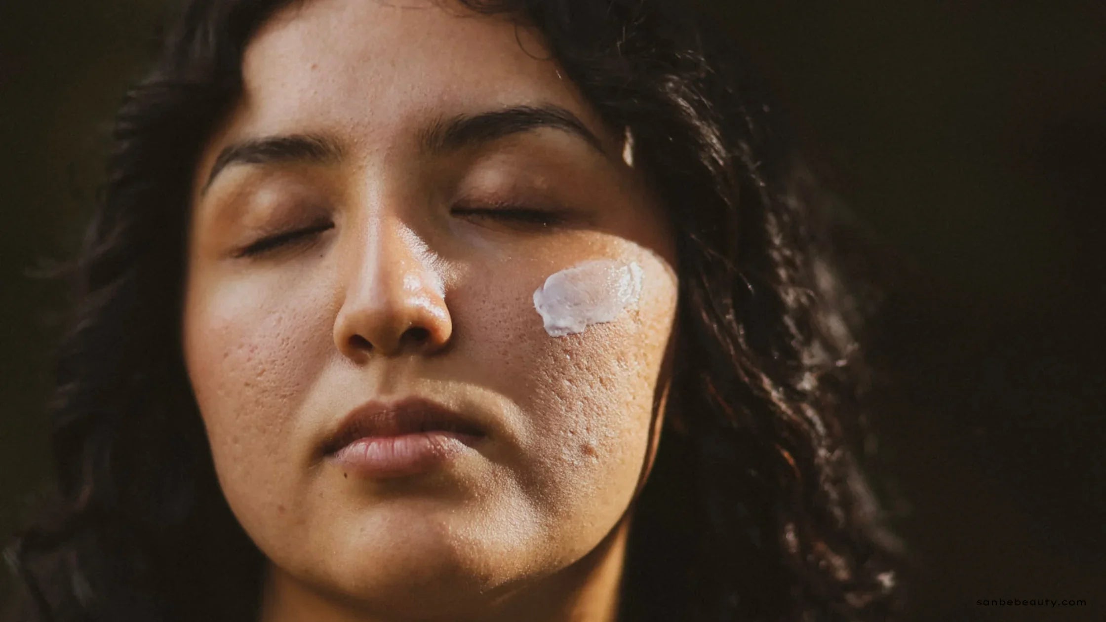 Close-up of a woman with her eyes closed applying a dollop of face cream to her cheek, illustrating the final moisturizing step in a skincare layering routine.